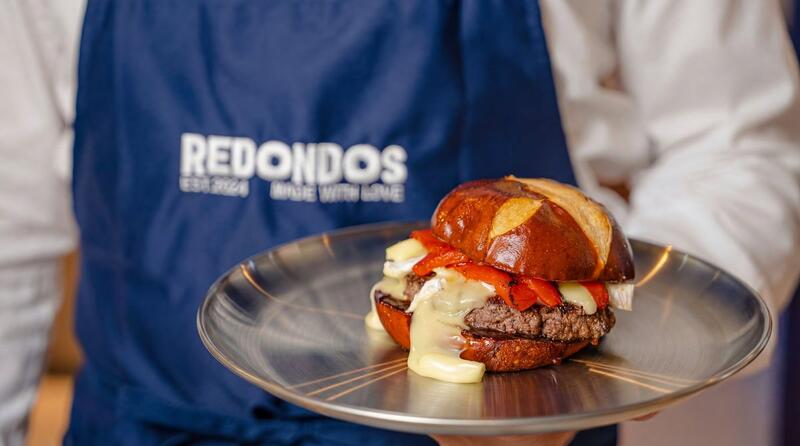 Signature Redondos smash burger with melted cheese on brioche bun served on silver plate by chef wearing REDONDOS apron